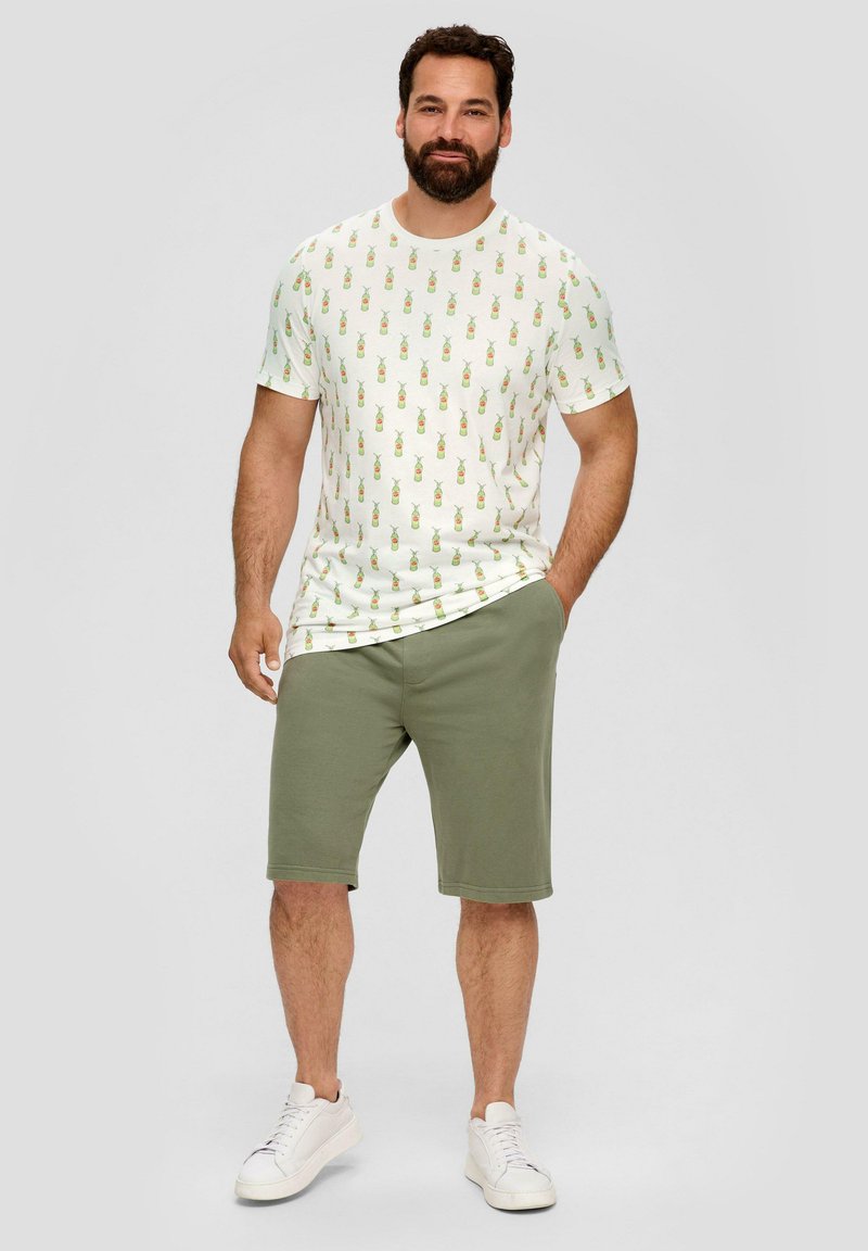 Witte T-shirt met groen ananaspatroon, gecombineerd met olijfgroene shorts. Model draagt witte sneakers. Casual kledingensemble.