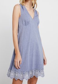 Blaues Chambray-Ärmelloses Kleid mit V-Ausschnitt, raffinierten Details und weißem besticktem Spitzenabschluss. Weicher Stoff und A-Linie-Silhouette.