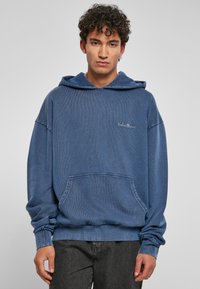 Felpa blu a coste con cappuccio e tasca frontale, caratterizzata da un logo ricamato sul petto e polsini elastici sulle maniche.