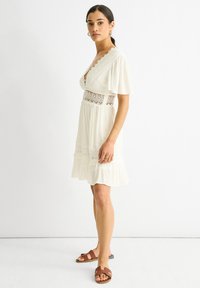 Robe blanche avec des détails en dentelle, manches courtes et jupe à volants. Présente un décolleté en V profond et une section centrale en crochet ouverte.
