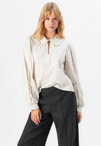 Lysfarget bluse med rund krage og sløyfedetalj. Har puffermer med scalloped detaljer og en nøkkelhullutringning.