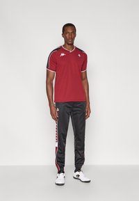 Czerwona sportowa koszulka z czarnymi akcentami i białymi logo, zestawiona z czarnymi spodniami z czerwono-białym paskiem wzdłuż boków.