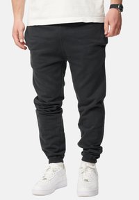 Schwarze Jogginghose mit lockerem Schnitt, elastischen Bündchen und Kordelzugbund. Aus weichem Stoff gefertigt, kombiniert mit weißen Turnschuhen.