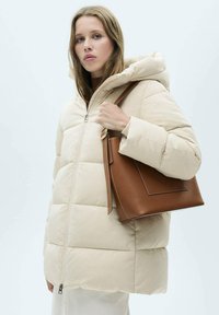Beige pufferjack met een ritssluiting aan de voorkant en een capuchon, met doorgestikte delen. Het model houdt een bruine tote bag vast met een gestructureerd ontwerp.