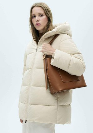 Beige pufferjack met een ritssluiting aan de voorkant en een capuchon, met doorgestikte delen. Het model houdt een bruine tote bag vast met een gestructureerd ontwerp.