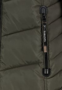 Donkergroen doorgestikt stof met een zwarte verticale rits en een zwart trekkoord met het label "SUBLEVEL" in witte tekst.