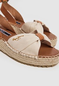 Espadrilės sandalai su smėlio spalvos kanvoso viršumi, surišto dizaino, rusvos odos akcentais ir su audinio juto padu, turinčiu tekstūruotą detales.