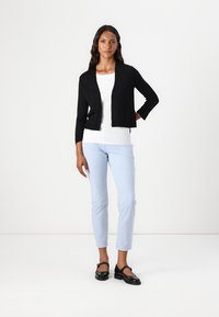 Zwarte cropped cardigan over een witte top, gecombineerd met lichtblauwe broeken. Model draagt zwarte schoenen. Glad stof en eenvoudig ontwerp.