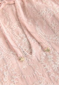 Tessuto rosa con motivi floreali ricamati bianchi, coulisse a cordoncino e punte metalliche dorate a forma di conchiglia.