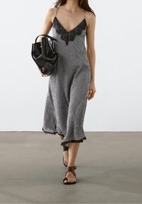 Vrouw die loopt in een zwart-wit geruite slipdress met kantafwerking, een zwarte handtas draagt en bruine kruisband sandalen aanheeft.