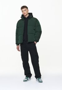 Chaqueta de plumas verde con cremallera en el frente y cuello alto, usada sobre un hoodie negro. Pantalones cargo negros y zapatillas verdes y blancas completan el atuendo.