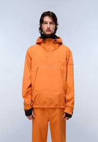 Ung person iført en orange hættetrøje og matchende bukser, der står med ansigtet fremad mod en ensartet lys baggrund.