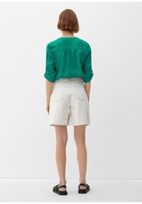 Grüne Leinenbluse mit aufgerollten Ärmeln, kombiniert mit beigefarbenen, maßgeschneiderten Shorts. Flache schwarze Sandalen runden das Outfit ab. Einfaches Design, ohne Muster.