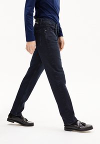 Jeans de mezclilla azul oscuro con un diseño de pierna recta, que cuentan con bolsillos traseros y una etiqueta de cinturón negra. Combinados con mocasines negros.