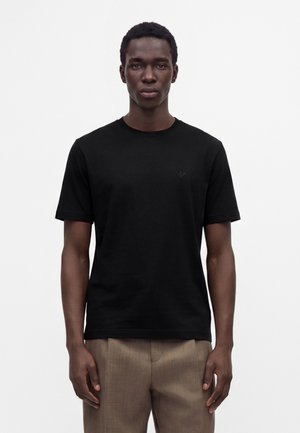 Ung mand iført en ensfarvet sort T-shirt med korte ærmer og lysebrune bukser, står foran en hvid baggrund og vender fremad.