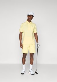 Polo de golf galben deschis, cu mâneci scurte, având un logo verde, asortat cu pantaloni scurți. Poartă o pălărie albă și pantofi de golf.
