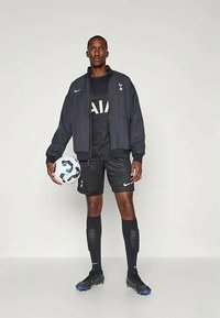 Black Nike jacket with vertical stripes, black jersey with "AIA," black shorts, and black cleats. Model holds a white soccer ball.