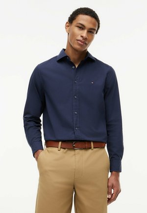 Mand iført en mørkeblå skjorte med knapper og beige bukser med brun bælte, stående med den ene hånd i lommen mod en hvid baggrund.