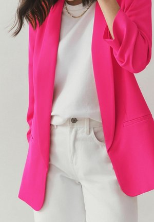 Femme portant un blazer rose vif sur une chemise blanche et un pantalon blanc, accessoirisée d'un collier chaîne en or.