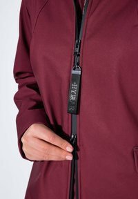 Chaqueta de abrigo burdeos con una textura suave, que presenta un cierre de cremallera y un tirador negro. El diseño incluye bolsillos laterales y un tejido suave.