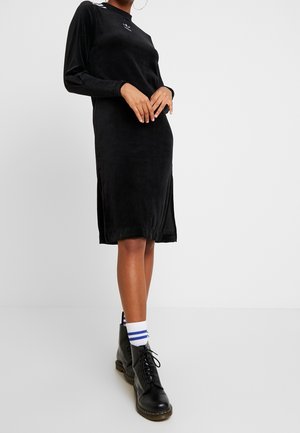 Personne portant une robe noire midi à manches longues, des chaussettes blanches à rayures bleues et des bottines à lacets noires, debout devant un fond blanc.