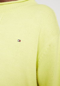 Jasnożółty sweter dziany o prążkowanej teksturze z małym logo w kolorach czerwonym, białym i niebieskim po lewej stronie klatki piersiowej.