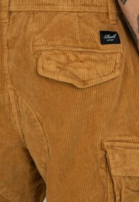 Corduroy overalls in medium brown, featuring vertical ribs, a back pocket with stitching detail, and a black label with white text.