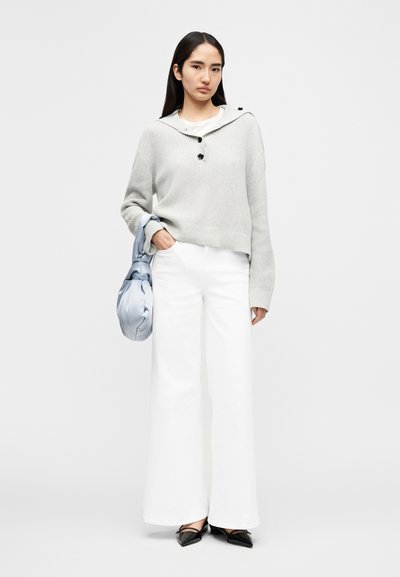 Ανοιχτό γκρι πλεκτό πουλόβερ με κουκούλα και λεπτομέρειες με κουμπιά, συνδυασμένο με λευκό παντελόνι φαρδιάς γραμμής. Κρατάει μια ανοιχτά μπλε μεταξωτή τσάντα και φοράει μαύρα φλατ παπούτσια.