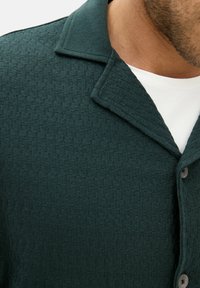 Vihreä neulepaita, jossa on teksturoitu kuvio, varustettuna käänteellä ja kahdella napilla, esitettynä valkoisen aluspaidan päällä.