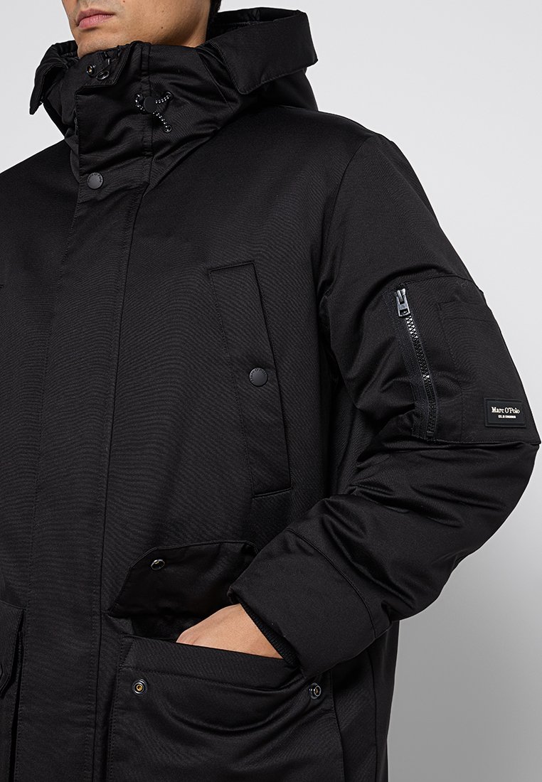 Veste extérieure noire avec une poche zippée sur le bras, de grandes poches à l'avant et un col haut. Fabriquée en matériau texturé et résistant à l'eau.