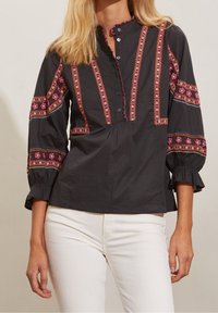 Chemise noire boutonnée avec broderies florales en rose, blanc et bleu, dotée d'un col et de manches à volants, assortie à un pantalon blanc.