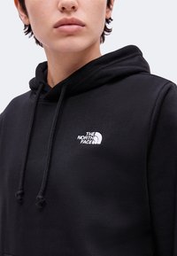 Personne portant un sweat à capuche noir avec cordons et un logo blanc The North Face sur la poitrine, visible des épaules aux lèvres.