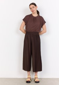 Brown short-sleeve top with pleat detail paired with brown wide-leg pleated pants and yellow sandals with fringe accent. Smooth texture.