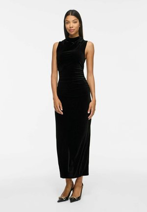 Femme debout portant une longue robe en velours noire sans manches et des escarpins noirs pointus avec un détail chaîne dorée sur fond blanc uni.