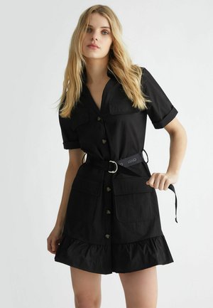 Abito nero a camicia con maniche corte, abbottonatura frontale, due tasche sul petto, orlo arricciato e vita fasciata con una cintura coordinata.
