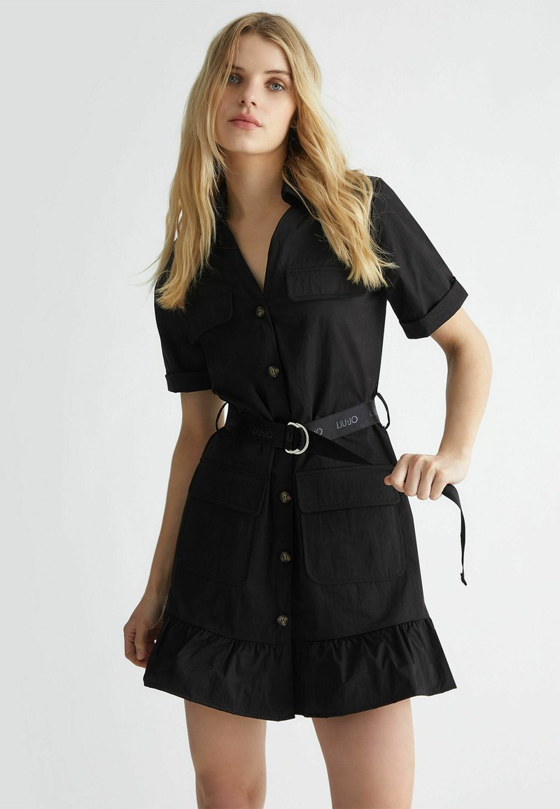 Abito nero a camicia con maniche corte, abbottonatura frontale, due tasche sul petto, orlo arricciato e vita fasciata con una cintura coordinata.