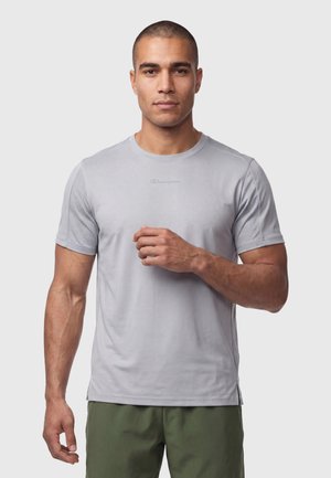 Mann mit kurzen Haaren, der ein hellgraues, kurzärmeliges Sportshirt und olivgrüne Shorts trägt und vor einem einfarbigen Hintergrund steht.
