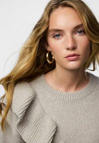 Svijetlosivi džemper rebraste teksture s naborima na ramenima. Model ima dugu, valovitu kosu i zlatne naušnice u obliku obruča.