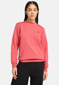 Sudadera rosa de tela suave con cuello redondo, mangas largas y un pequeño logo en el lado izquierdo del pecho. Combinada con pantalones negros.