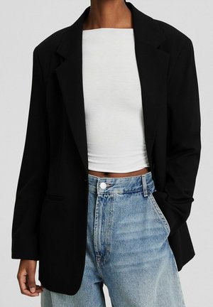 Sort oversized blazer med klippede revers, enkelt knappelukning og to frontlommer, parret med en hvid cropped top og lys blå jeans.