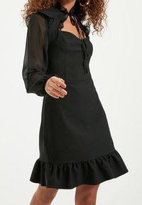 Svart klänning med en figursydd bodice och volangdetaljer, med genomskinliga långa ärmar och ett band i halsen. Slät textur.