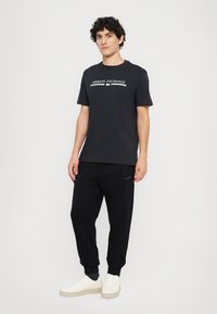 Svart t-skjorte med kort erm og "Armani Exchange"-logo, sammen med svarte smale joggebukser og hvite sneakers. Enkel design og uformell passform.