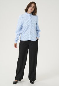 Blouse bleu clair avec un motif zigzag et un plastron à volants, assortie à un pantalon noir taille haute à jambes larges et des chaussures à talons noires pointues.