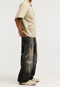 Beige kortärmad skjorta med ficka, kombinerad med överdimensionerade svarta jeans med graffiti-liknande mönster och lätt slitage. Sneakers fulländar looken.