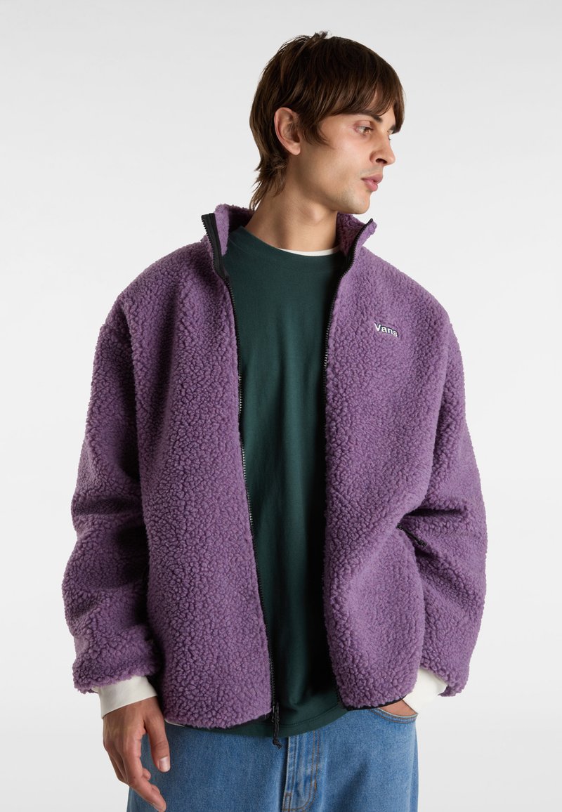 Lila Fleecejacke mit durchgehendem Reißverschluss, gerippten Bündchen und Seitentaschen, getragen über einem grünen Shirt; strukturiertes, weiches Material.