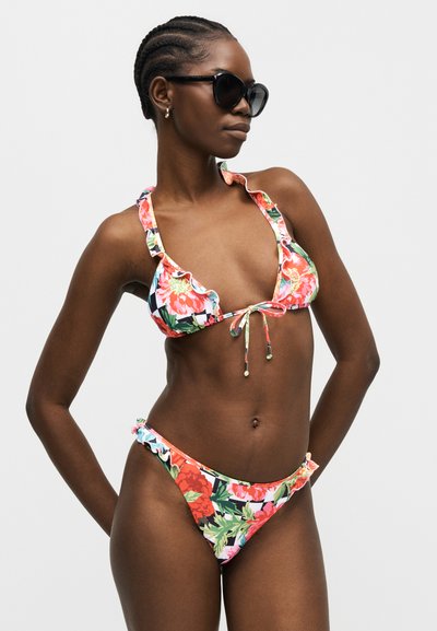 Γυναίκα με φλοράλ μπικίνι με βολάν στα άκρα και μαύρα γυαλιά ηλίου, στέκεται με τα χέρια στη μέση μπροστά σε απλό γκρι φόντο.