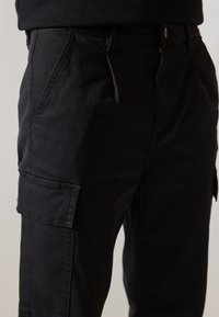 Schwarze Cargo-Hosen mit Seitentaschen, aus einem robusten Baumwollmix gefertigt. Sie verfügen über eine lässige Passform und eine leichte Textur. Mit Knopf- und Reißverschluss.