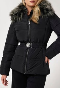 Juodas paminkštintas džemperis su kailiniu kapišonu. Yra sidabrinė sagtis, elastinė talija ir užtrauktukas. Tekstūruotas audinys su horizontaliais siūlais.