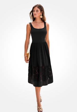 Mujer con vestido negro sin mangas con borde de encaje, pendientes dorados, collares en capas, llevando un bolso dorado, de pie frente a un fondo blanco.