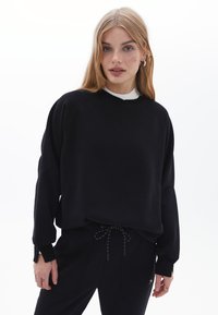 Felpa nera con maniche raglan, realizzata in tessuto morbido. Presenta un collo rotondo e polsini con texture a costine, abbinata a pantaloni neri con cordino.
